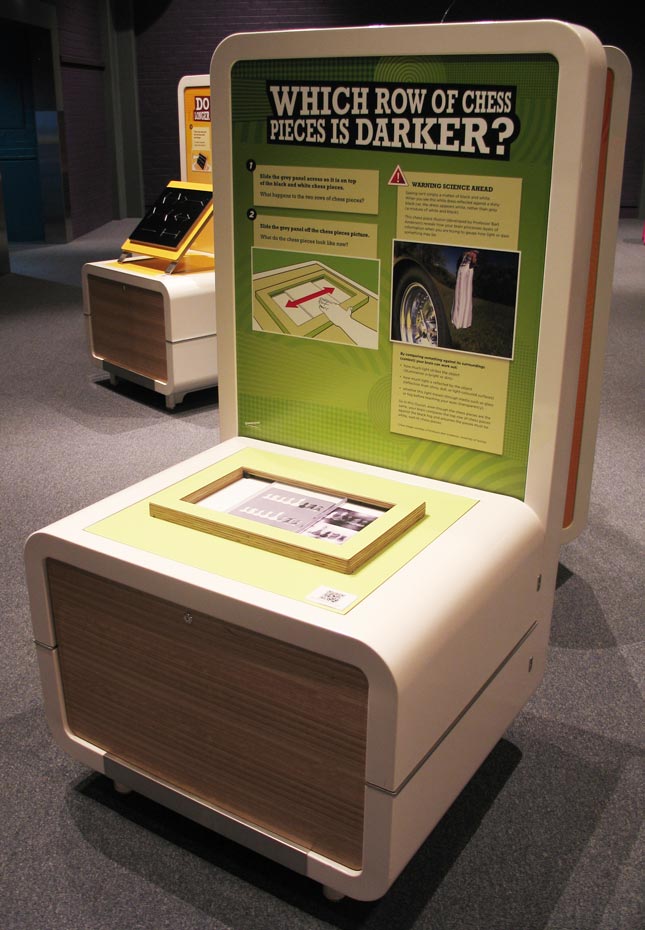 A cream and green exhibit table and information panel with the title 'Which row of chess pieces is darker?'.