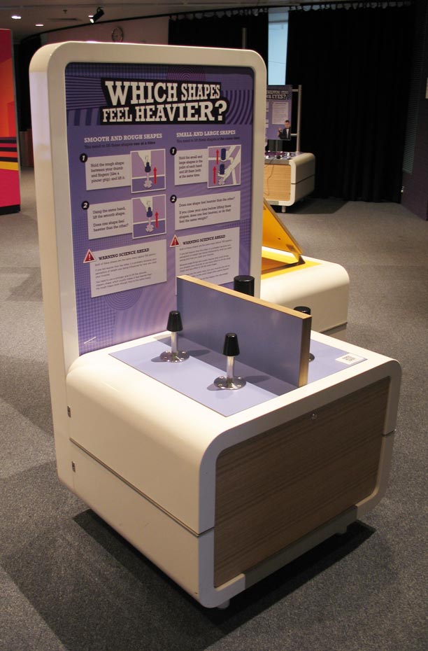 Lift two different sets of shapes or weights. One pair of shapes shows the texture-weight illusion, while the other pair of shapes shows the size-weight illusion. A cream and purple coloured exhibit table and headboard with the text 'Which shapes feel heavier?'. On the table top sits a divider board with four black cylindrical objects.