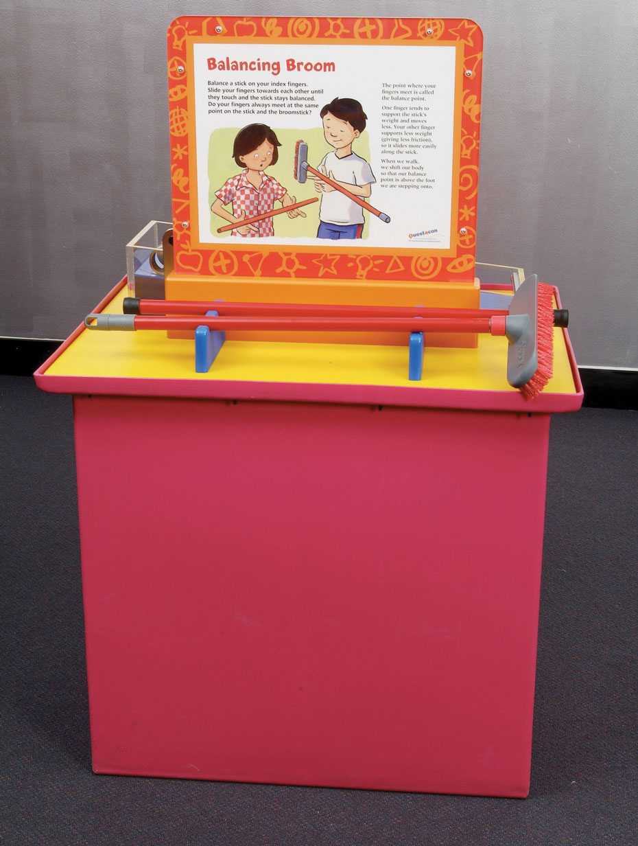 Balance the broom on your fingers and compare the balance point on a broom stick and a normal stick. A red exhibit table with a yellow top and a vetical orange and white information panel. In front of the panel is a red and grey broom.
