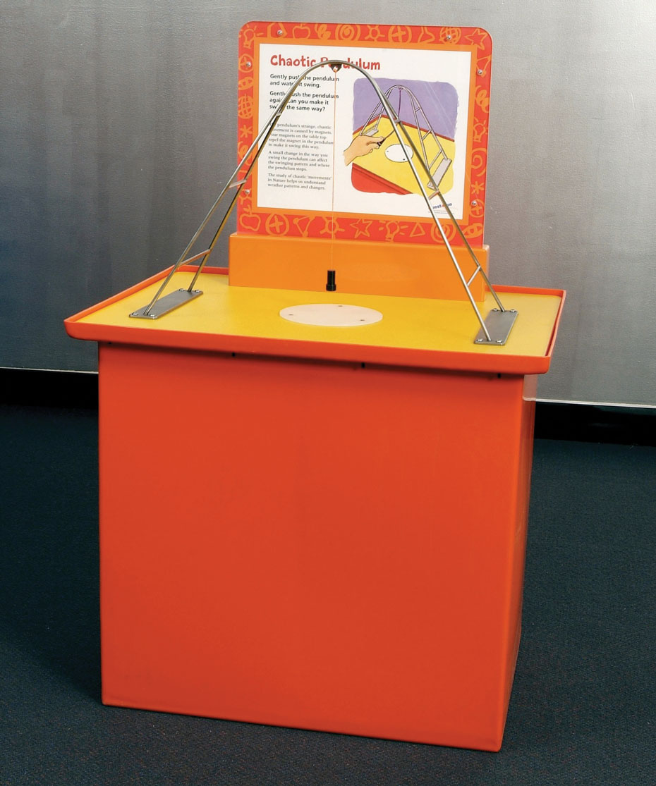 Push a magnetic pendulum and watch it swing chaotically over a metal plate. An orange and yellow exhibit table with matching information panel at the back and on the top is a steel arch with a string and black weight hanging down from the apex of the arch.