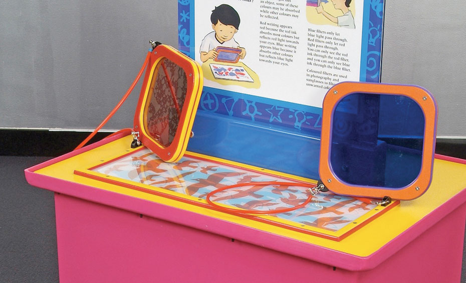 Look at a picture through a red filter, then a blue filter to observe how light wavelengths can be absorbed or allowed to pass through a filter. A pink and yellow exhibit table, with matching white and blue information panel on top. Sitting on top of the table are two square orange frames. One frame has a red see through filter in the centre, the other a blue one.