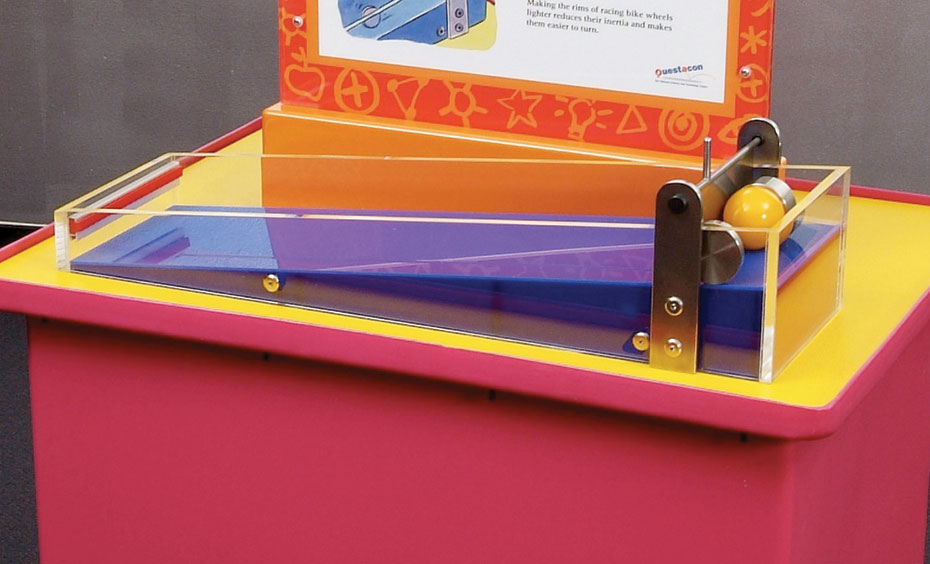 Have a rolling race between a ring, flat disc and ball to explore rotational inertia. Stand all shapes on their side behind the gate. Raise the gate to start them rolling at the same time. A red and yellow exhibit table, with an orange and white information panel on top. On the table top sits a purple long wedge contained in clear perspex and has a yellow ball at the high end contained by a steel gate.