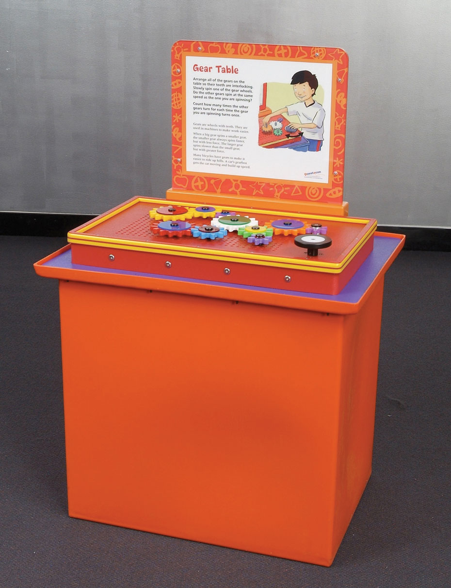 Different sized gears turn at different speeds and force to generate mechanical advantage. An orange, purple, red and yellow exhibit table with a white and orange information panel at the back. On top of the table are a series of colourful gears.