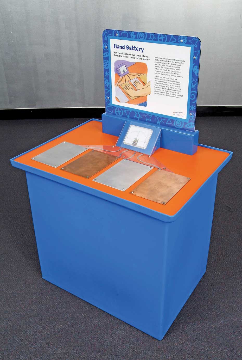Electric fields can be created when a circuit is closed in a loop formation. A blue and orange exhibit table with a white and blue information panel at the back. On the top of the table are four metal rectangular pads connected to a voltage meter.