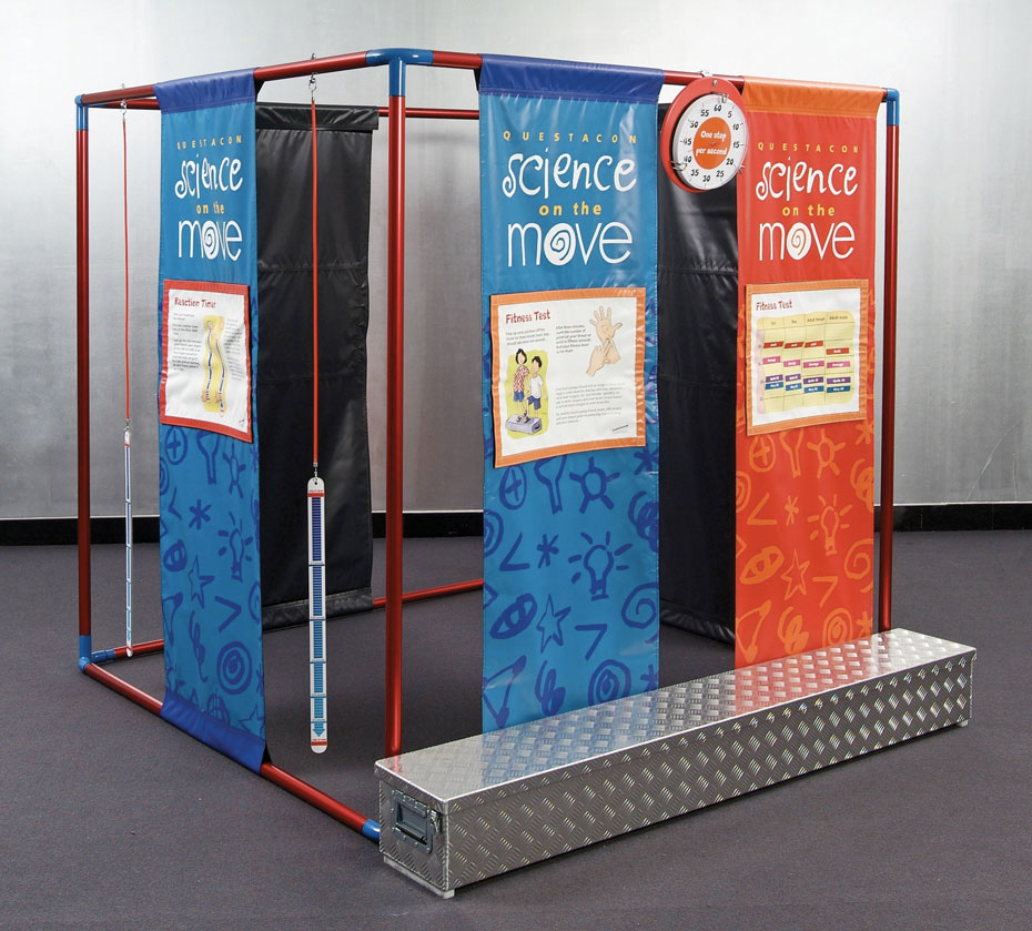 One person drops a reaction stick, so a second person must catch and measure their reaction time. A large red and blue cube frame that has blue and orange banners running from top to bottom with different information on each. In front on the grey carpet is a long checkerplate steel box.