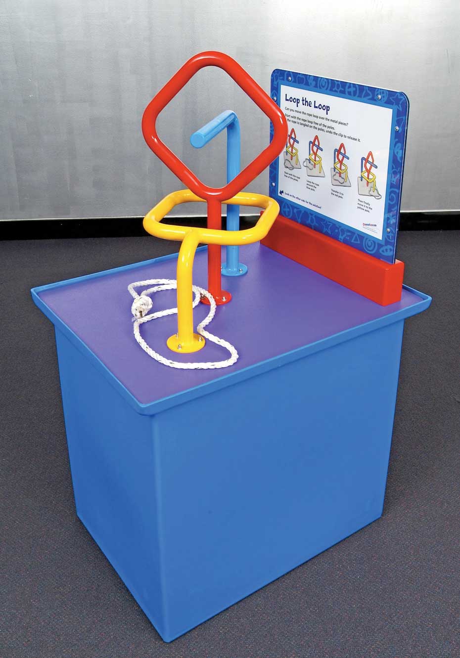 Topology puzzles explore patterns in space. A blue exhibit table with a blue, white and red information panel on top. On the top of the table intertwined red, blue and yellow steel bars, with a white rope with a knot tied in it, that is around the yellow bar.