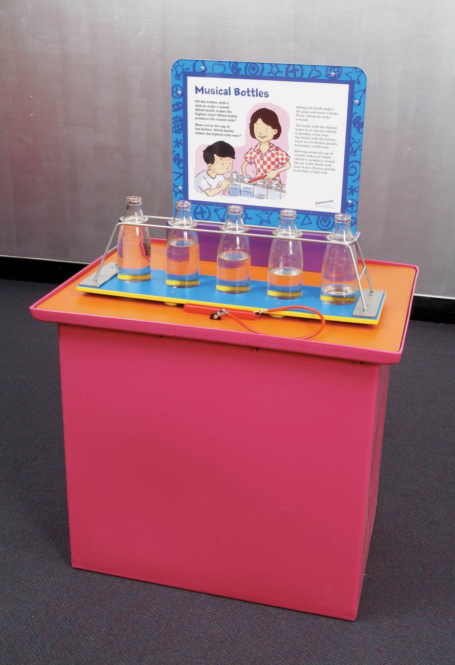 Hit bottles filled with varying levels of water to play a tune. A red and orange exhibit table, with a white and blue information panel at the back. On top of the table are 5 milk bottles filled with different levels of water.