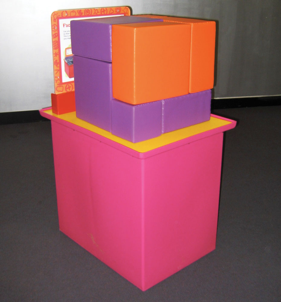 In this puzzle, pack nine small parcels so they fit efficiently into one large container. A pink and yellow exhibit table, with a white and orange information panel at the back. On the table sits purple and orange soft square shapes that all fit together to form a cube.