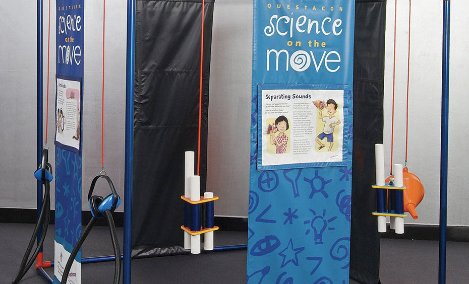 Listen to ambient sounds through plastic funnels that channel sound waves to the ear. a blue, black and white cube structure that has vinyl blue and white information panels from top to bottom with spaces in between. There are also different sound exibits hanging from orange cord from the cube structure.