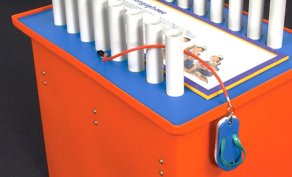 Use the rubber shoe to strike the open ended pipes and play a musical tune. An orange and blue exhibit table with a white information panel laying flat on the top, two rows of 20 millimetre pvc piping rising up from the table top, and a blue thong attached to an orange cord hanging over the table side.
