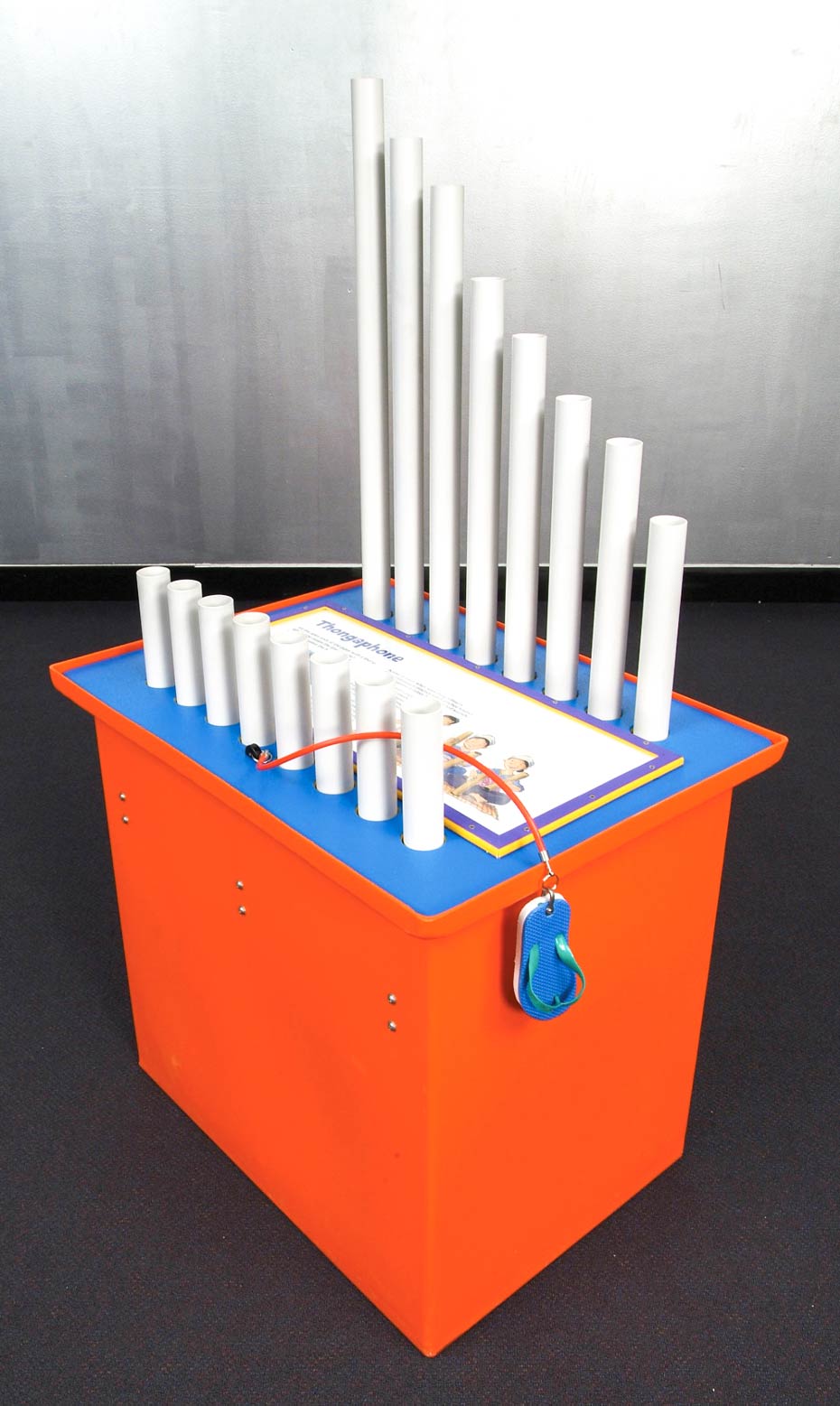 Air vibrates at different frequencies to generate low or high pitched sounds. An orange and blue exhibit table with a white information panel laying flat on the top, two rows of 20 millimetre pvc piping rising up from the table top, and a blue thong attached to an orange cord hanging over the table side.