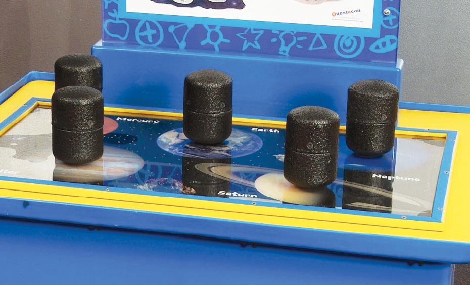 Lift pods that represent the same mass, but different weights on certain planets due to gravity. A blue and yellow exhibit table, with a white and blue informationa panel at the back. On the table top sits 4 black weights.