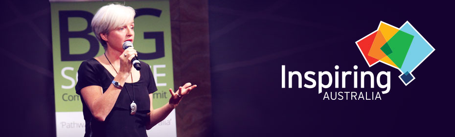 A woman with blond hair, wearing a black t-shirt and holding a microphone is standing in front of a green and white sign. To the right is the logo for Inspiring Australia