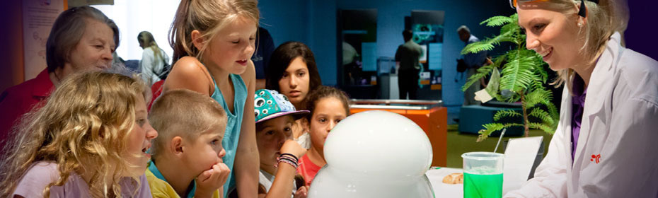 A young woman dressed in a lab coat is demonstrating how bubles are made to a bunch of young school kids.