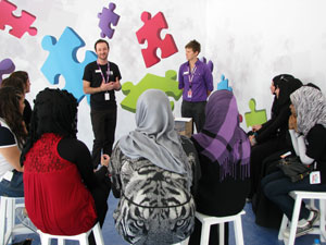 A group of women in middle eastern dress are sitting in a semi-circle listening to a man and woman talking.
