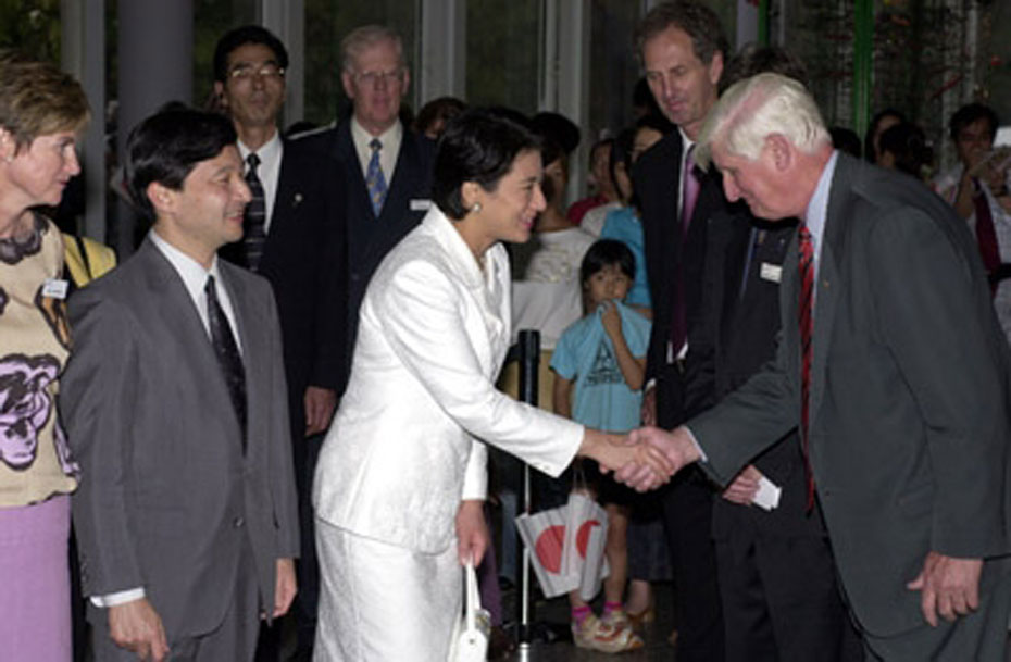 This photo shows Mike Gore, former Questacon Director, shaking hands with Crown Princess Masako of Japan at an official ceremony. Crown Prince Naruhito stands beside the Princess. Dr Gores nods respectfully. The royal couple are smiling. In the background, children and dignitaries look on.