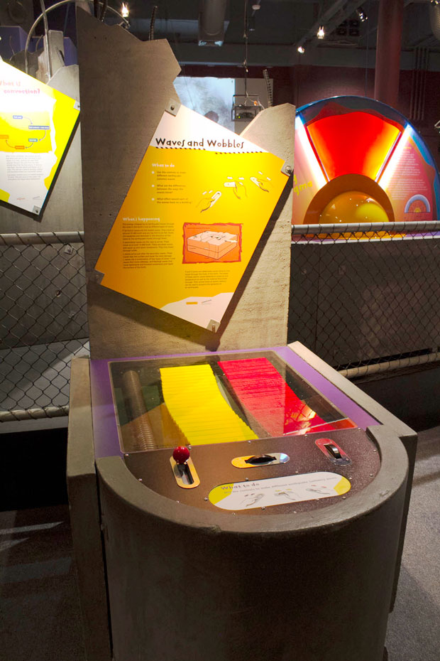 Use the controls to make different earthquake (seismic) waves move along the rods or spring. A yellow and red series of horizontal rods under a glass display case. The horizontal component of the case has a yellow information panel.