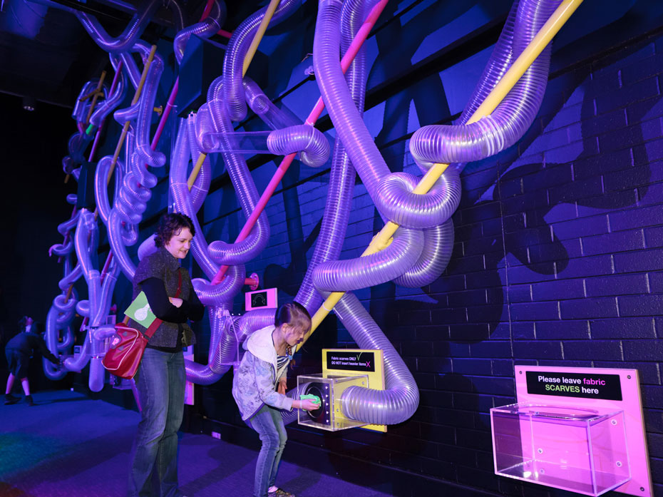 Insert one or more fabric scarves into an inlet box and watch how the scarves fly through a labyrinth of air tubes. A young girl placing a green scarf in a perspex box that is connect to a series of large air tubes mounted on a purple/blue wall. A woman watches on behind her.