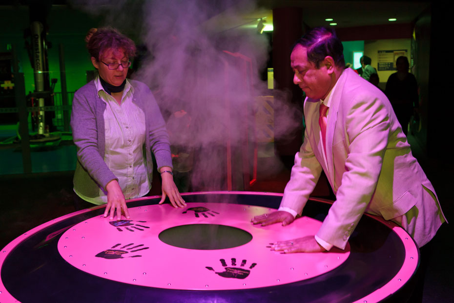 If everyone pushes down on one side, which direction will the vortex go? A woman and man pressing down with both sets of hands on a round table. There is smoke rising from the centre of the table, and hand prints painted on the table top surface.