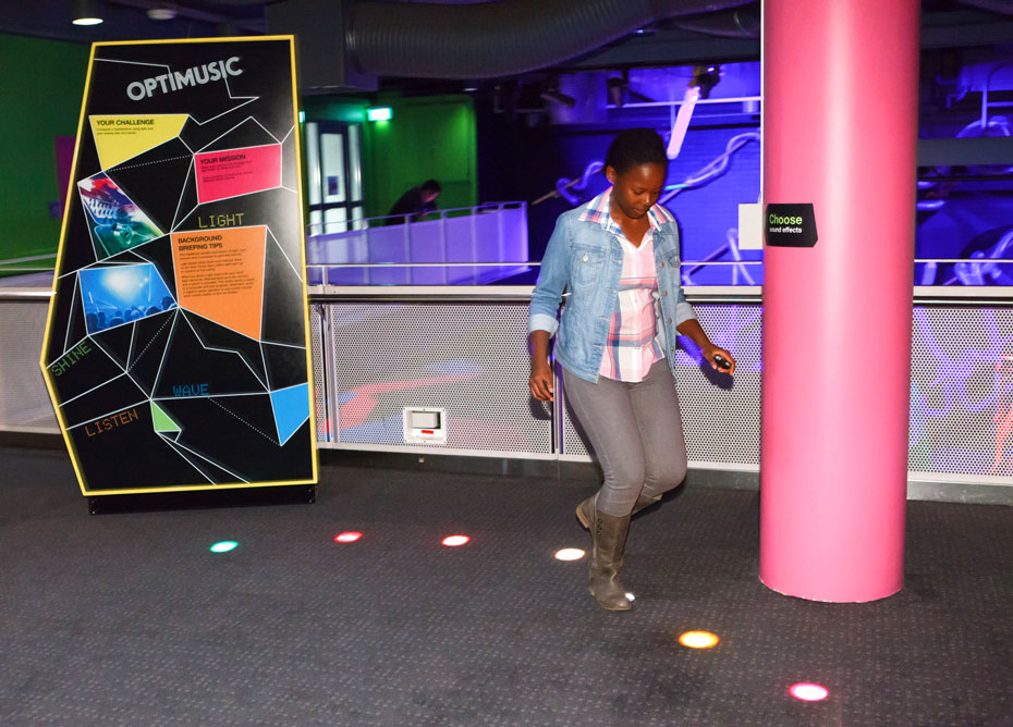 Press a button to choose different sound effects, then block beams of coloured light by walking through the beams, or waving your feet into the lights to generate a sound. A multi coloured sign with the words 'Optimusic' at the top, along side a carpeted area with a pink structural pole, and different coloured floor lights in a curved line. A teenage girl is walking on the lights.