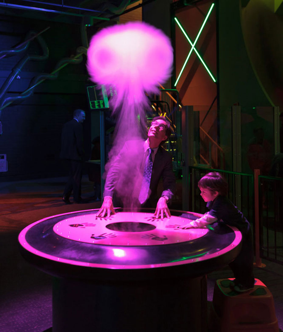 Push down on the drum’s collar. Watch how a vortex ring floats up towards the ceiling and make the ribbons on the ceiling curl and sway in response. A man and child with both hands on a round table watch as a jellyfish shape cloud rises from the centre of the table.