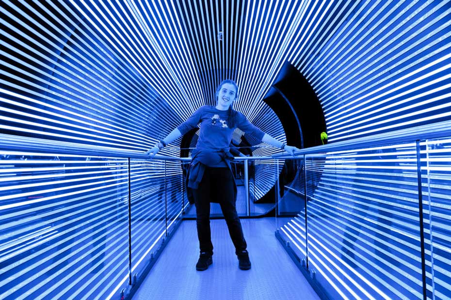 A loss of balance can be created by fooling your sensory inputs. A teenage girl holds onto hand rails in a large cylinder lined with blue and white led lights. She is leaning to the right.