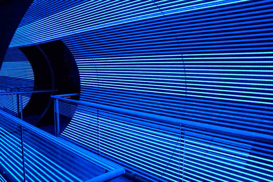 This Rototron tunnel turns LEDs on and off to generate the visual illusion of movement. White and blue led lights line the inside of a large cylinder you can walk through. There is also hand railing in the photo.