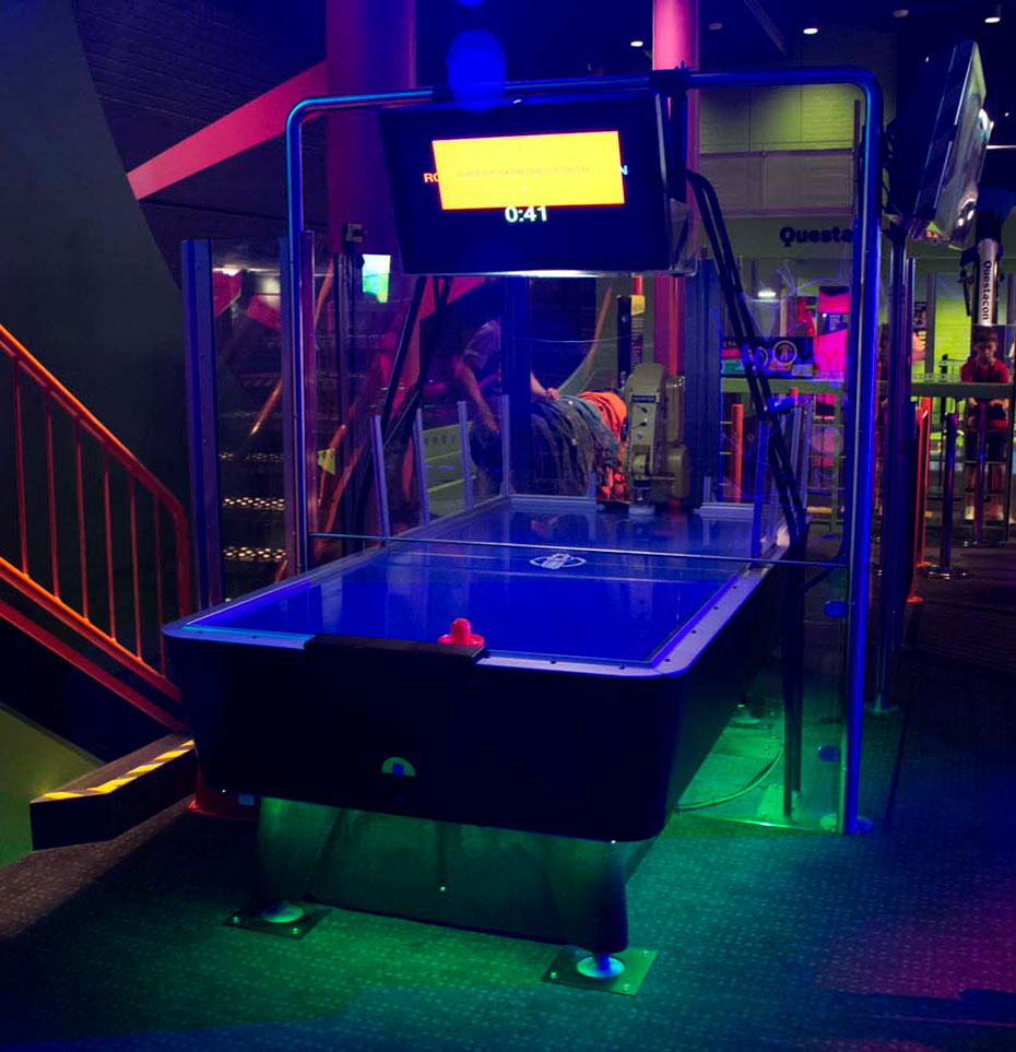 Compete against a robotic arm in a game of air hockey. A screen displays the number of points (goals) won by the humans or the robot during a game. A blue hockey table with a tv screen above it and perspex glass enclosing the a robot arm at the back.