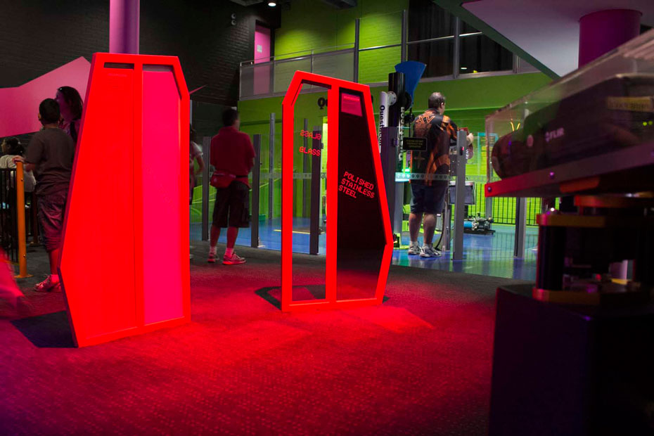 Find a panel of material to hide behind to avoid being detected by the thermal camera. The video screens show what the thermal camera ‘sees’. A black thermal camera enclosed in a protective perspex box, pointing at two red upright display boards with people in the background