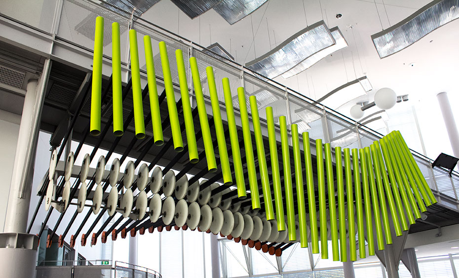 Is there a constant wave pattern, or does the wave slowly change over time? A row of vertical green pipes in a wave formation, sit under a overhead walkway. They both sit within a large foyer.