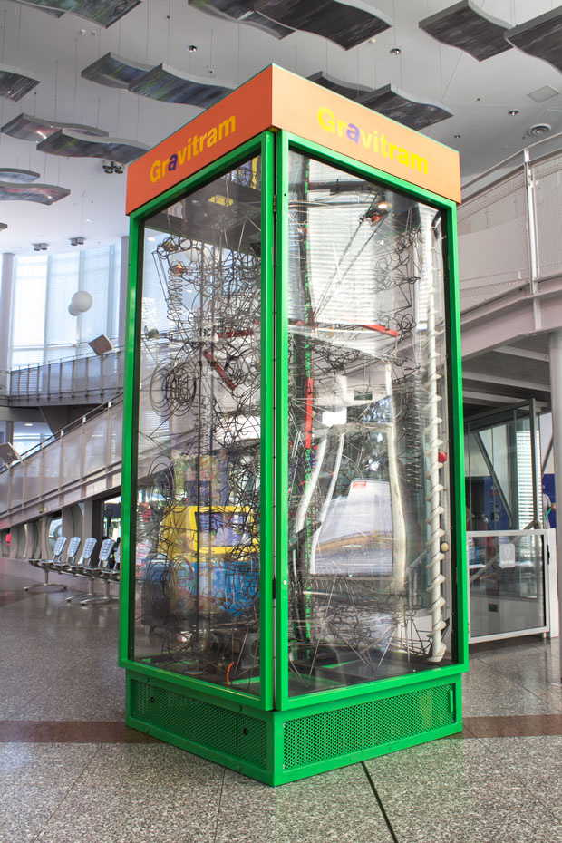 Kinetic sculptures lift, roll and drop billiard balls around various pathways. A large green steel and glass display case sits within a white foyer. Within the display case is a series of wire pathways and billard balls.