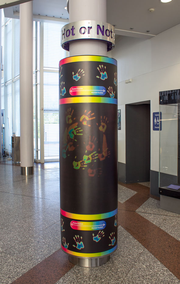 Place your hands on the pillar, then lift your hands to see a coloured imprint. A large grey and black pillar with a sign saying 'Hot or Not' above it. The black pillar has rainbow colours in horizontal bands top and bottom, while there is also colourful hand prints inbetween the bands on the black areas.