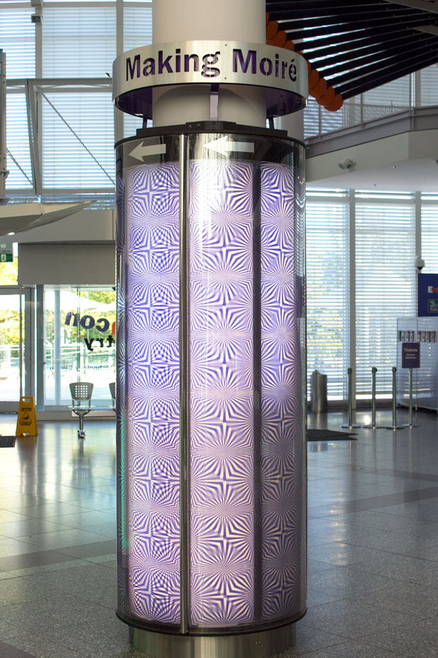 Spin the outer sheath of the pillar and watch how the patterns form. A blue and white pillar in a glass and white foyer, with a sign at the top saying 'Making Moire'.