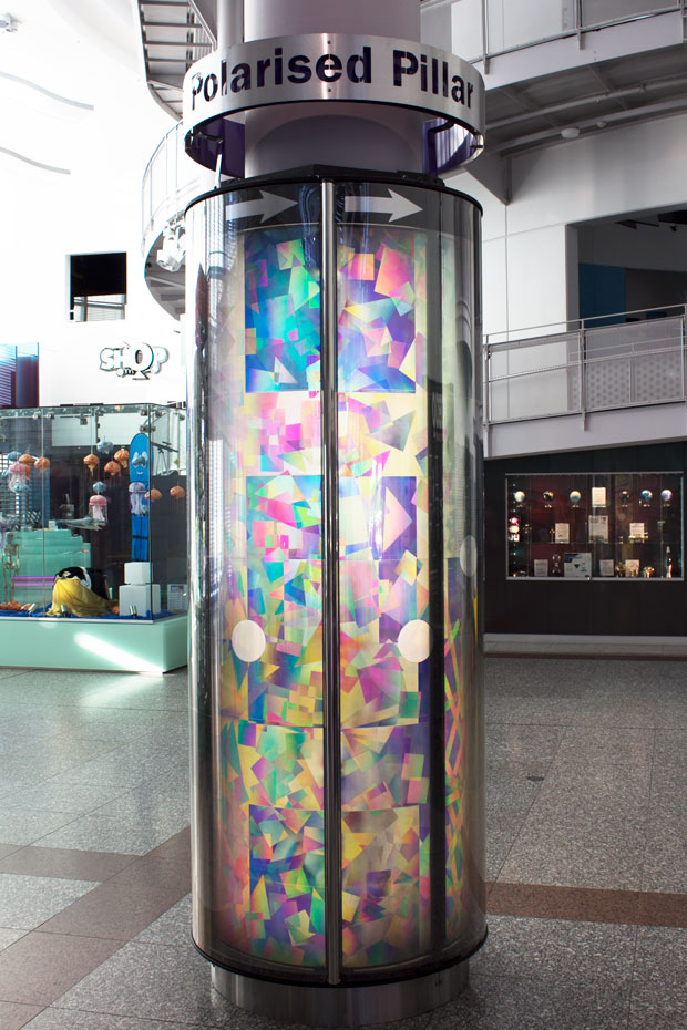 Spin the outer sheath of the pillar and watch how the polarising filter blocks or filters light. A large pilar display with different geometric shapes, sizes and colours. The pillar sits in a large foyer.