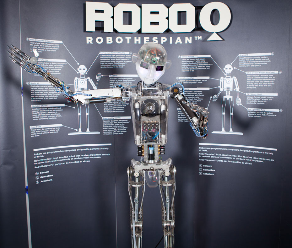 Use a nearby touch screen to control RoboQ's actions and speech. A robot with it's arms spread outwards, and a large grey and white information panel behind it with the title 'Robo Q Robothespian'.