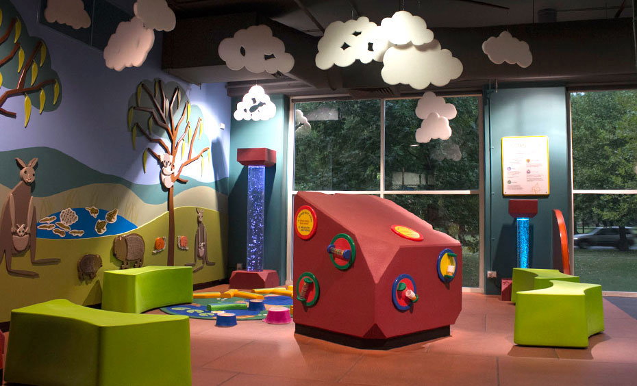 Several exhibits in Sensory Space encourage young children to match sights, sounds and textures, as they are encouraged to turn discs or watch and listen to the bubble towers. A brightly coloured play room with a billabong scene on one wall, cut out clouds hanging from the ceiling, and an array of play objects in the picture.