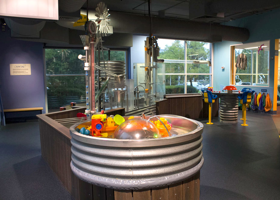 Playing with water improves a child’s ability to plan, measure and predict events (such as whether something will float or sink). An overview of the exhibit, with a steel water tub in the front, a wind water pump in the background and a long bench running between them.