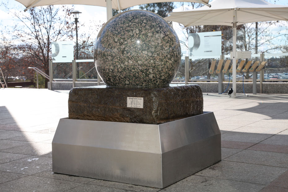 Push on the large granite ball to make it spin within its cradle. A large granite stone ball sitting on a large slab of stone, which intern sits within a stainless steel bin. Water flows from under the granite and over the stone to the bin.