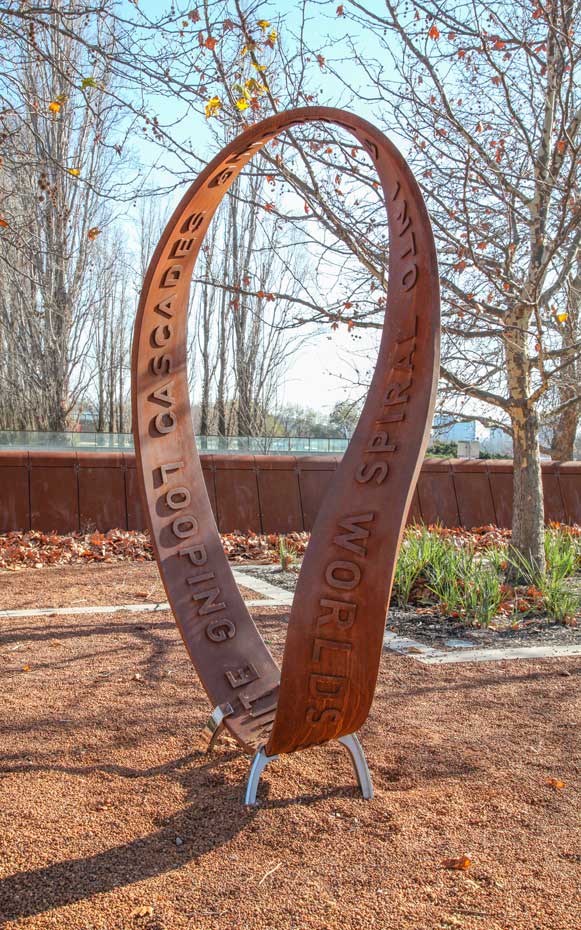 Select one spot on the sculpture and imagine tracing a toy car running around the track and returning to the starting position. A rusted continuous steel loop sculpture with the raised words 'looping cascades' and 'into sprial worlds' on the surface of the strip. The sculpture sits on a brown graveled garden area, with deciduous trees in the background.