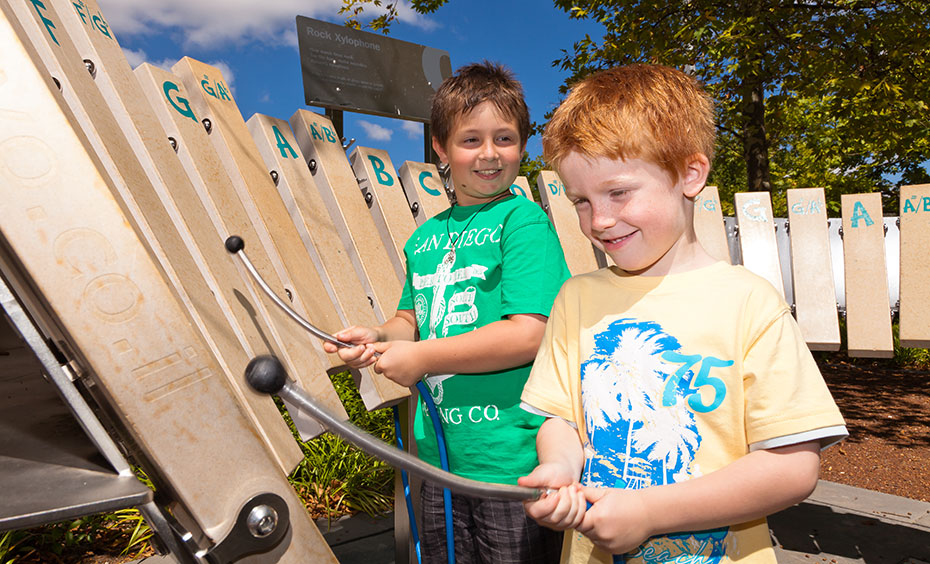 Gently tap each limestone bar to hear a different pitched sound and play your tunes of choice. Two young boys each with a metal mallet and rubber mallet are hitting semi-vertical stones notes arranged in a keyboard arrangement.