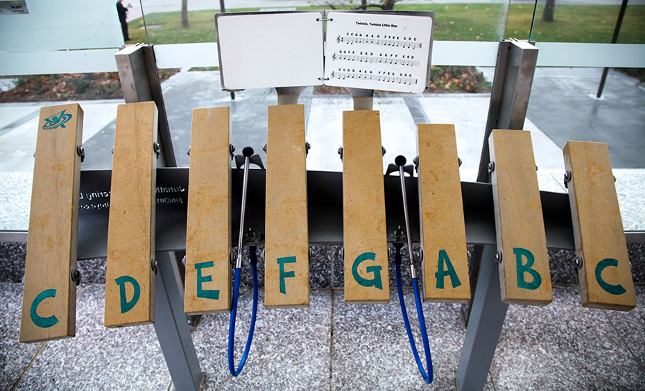 Gently tap each limestone bar to hear a different pitched sound and play your tunes of choice. Eight limestone blocks layout as a keyboard on a metal stand, with two blue plastic and metal mallets attached. Behind the keyboard is sheet music for 'Twinkle, Twinkle Little Star' with the whole exhibit sitting on a granite and glassed area, with gardens in the background.