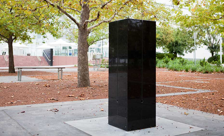 While solid objects vibrate to create sound, air gaps help to generate louder sounds. A rectangular black granite pilar about 1500 milimeters heigh, with for slits running from across the top to three quarters of the way down the sides. The pilar sits on concrete and is located in a garden environment.