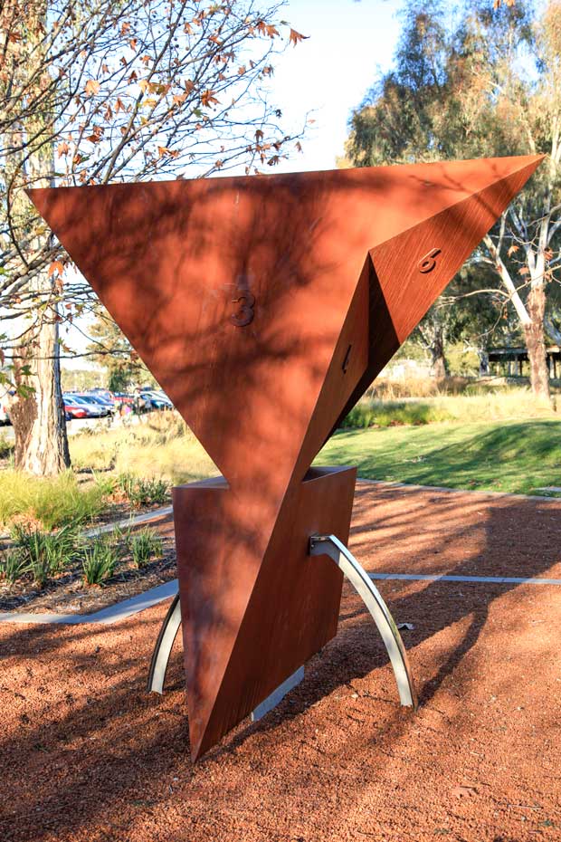 A side view of the Szilassi Polyhedron showing the different face shapes. A rusted metal sculpture with six faces located in a garden environment.