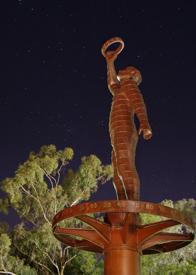 The Astronomer at night. Why is the astronomer holding a ring? A night time photo of a rusted metal sculpture of a man holding a ring up to the sky with one outstretched arm. There are gum trees lit up in the background and the sky is black with thousands of stars visible.