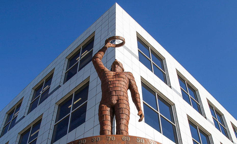 An artistic sculpture by Tim Wetherell, representing parallax to measure astronomical bodies and recovery from the 2003 Canberra Bushfires. A rusted metal sculpture of a man holding a ring up to the sky with one outstretched arm. Behind him is the corner of a large white building with dark windows and a cloudless blue sky.