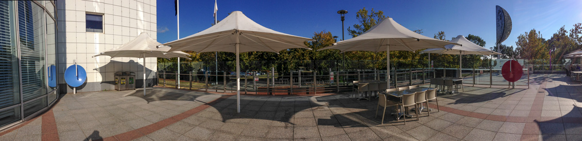 One person whispers into the focal point of one whispering dish, while a second person stands listening at the focal point of the second dish. A panoramic photo of a courtyard with large umbrellas, tables and chairs and glass safetly railing. To the far left is a white wall and window with a large vertical blue dish, and to the right is a large red dish identical to the blue one. There are trees in the background.