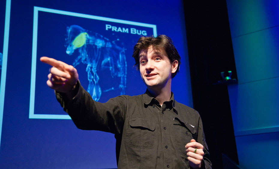 A man wearing a collared shirt is pointing and sanding in front of a screen that has a blueprint of a bug on it.