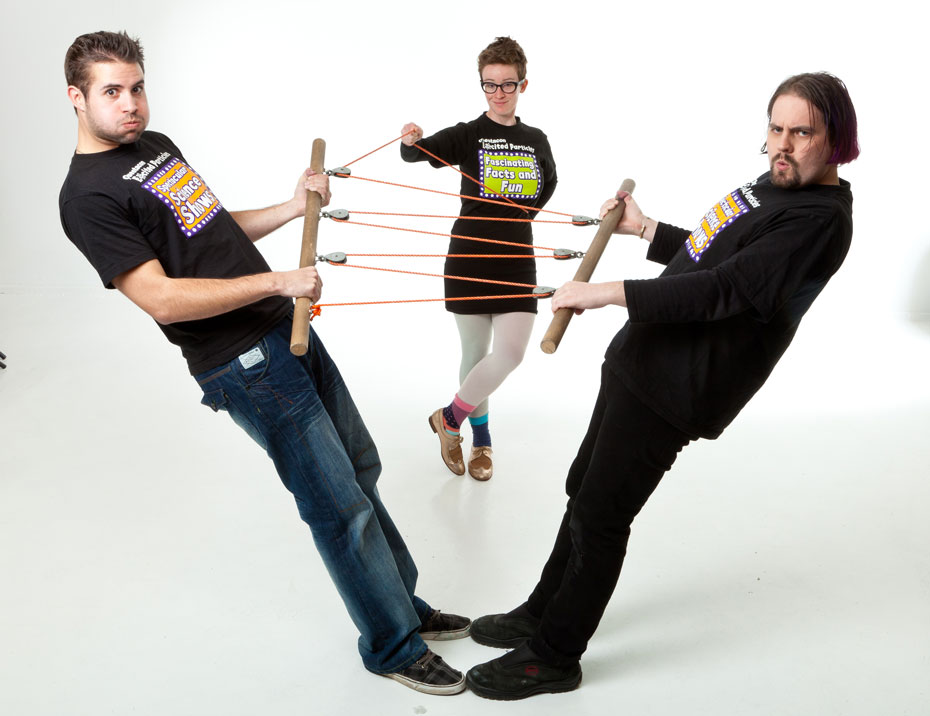 Two men leaning away from one another each have a wooden pole and pulleys and rope attached to each. A woman is standing at the centre of picture in between them also holding onto one of the ropes.