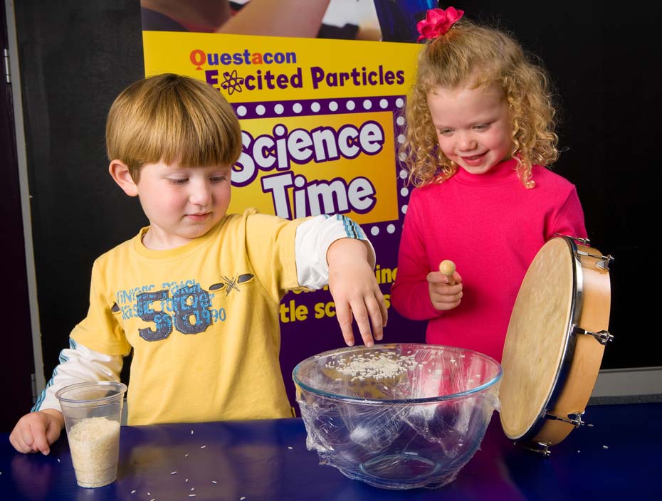 A young girl and boy playing with a tambourine and a large clear bowl with plastic film stretch across the top and rice sitting on this plastic film.