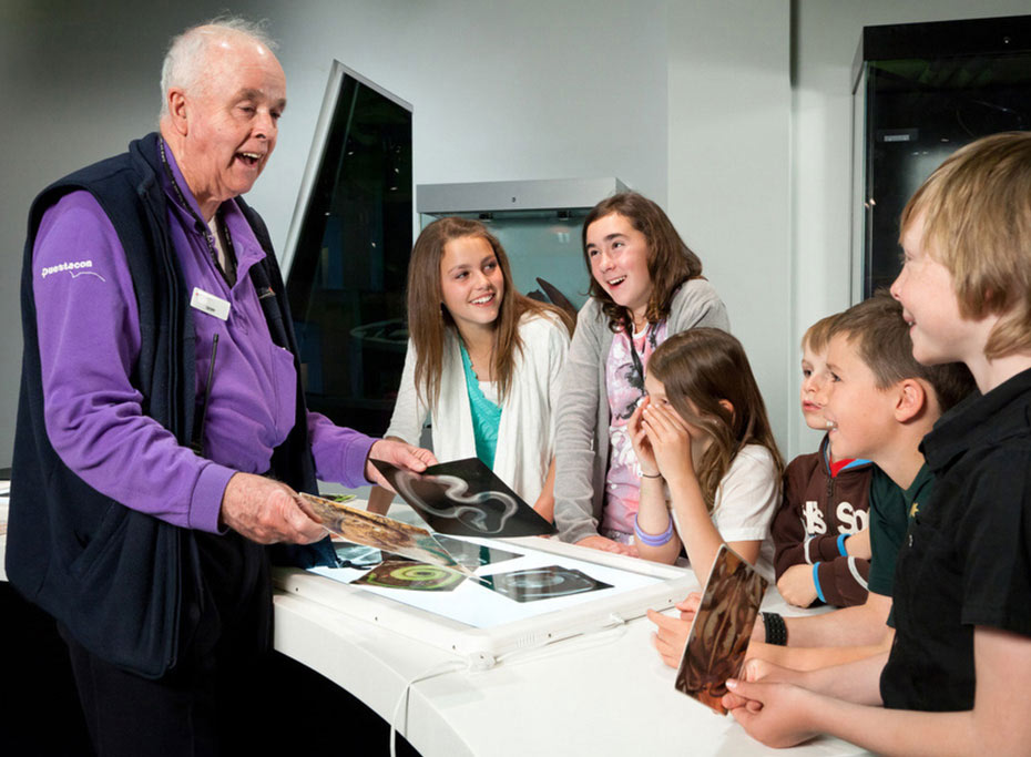 A group of young children and teenagers are interacting with a elderly man who is showing them various photographs.